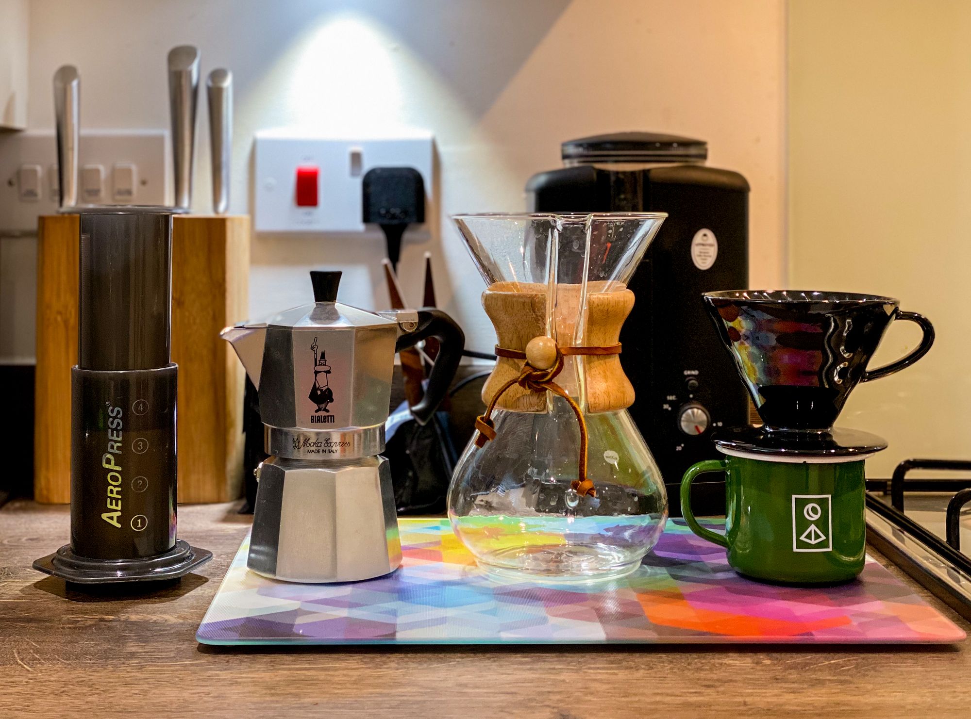 An Aeropress, Moka Pot, Chemex dripper and a black ceramic V60 on top of a green enamel mug, all placed on a kitchen counter top