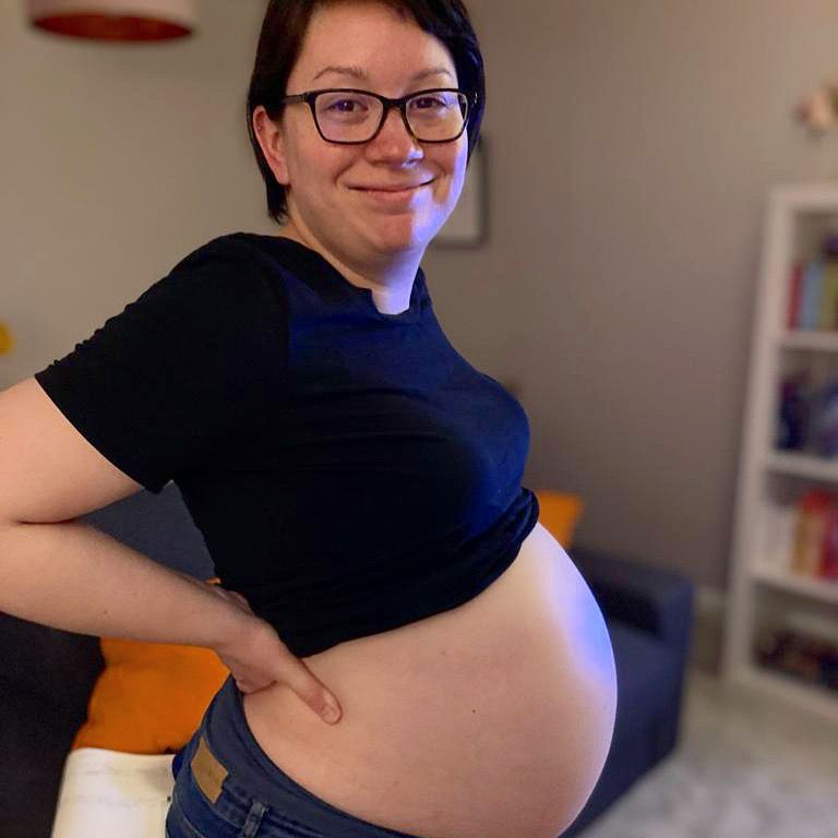 Me, Jess Darnes - a thirty-something year old white woman with short dark hair and glasses - standing with my baby bump out, smiling, a few days before giving birth