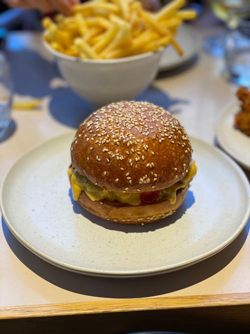 A bacon cheeseburger on a beige plate with a big bowl of fries in the background